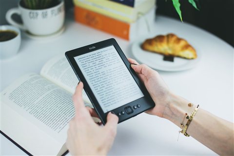 Person holding a Kindle e-reader next to a coffee cup while it charges via cable Person holding a Kindle e-reader next to a coffee cup while it charges via cable