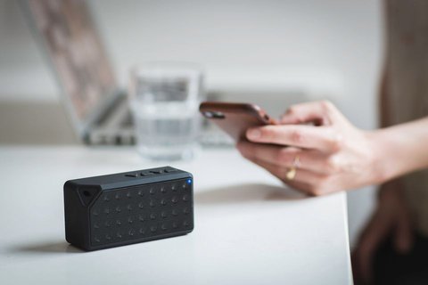 Hand holding phone next to small black portable Bluetooth speaker on desk Hand holding phone next to small black portable Bluetooth speaker on desk