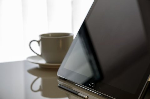 Tablet standing upright on a glass table beside a coffee cup and stylus pen Tablet standing upright on a glass table beside a coffee cup and stylus pen