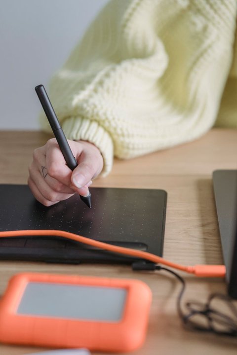 Close up of a stylus pen resting on a black drawing tablet next to a laptop