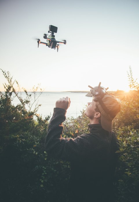 FPV racing drone hovering outdoors while pilot wears goggles nearby FPV racing drone hovering outdoors while pilot wears goggles nearby