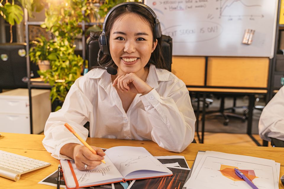 Smiling woman wearing a wireless headset with boom mic taking notes at a desk Smiling woman wearing a wireless headset with boom mic taking notes at a desk